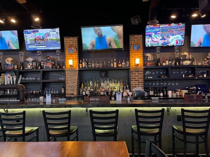 Empty bar with barstools, liquor bottles on shelves, sports memorabilia, and three TV screens displaying sports, viewed from across a wooden table.