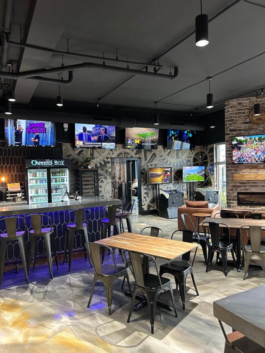 A modern sports bar with multiple TVs, a bar counter with high stools, tables with chairs, and a refrigerator stocked with drinks.