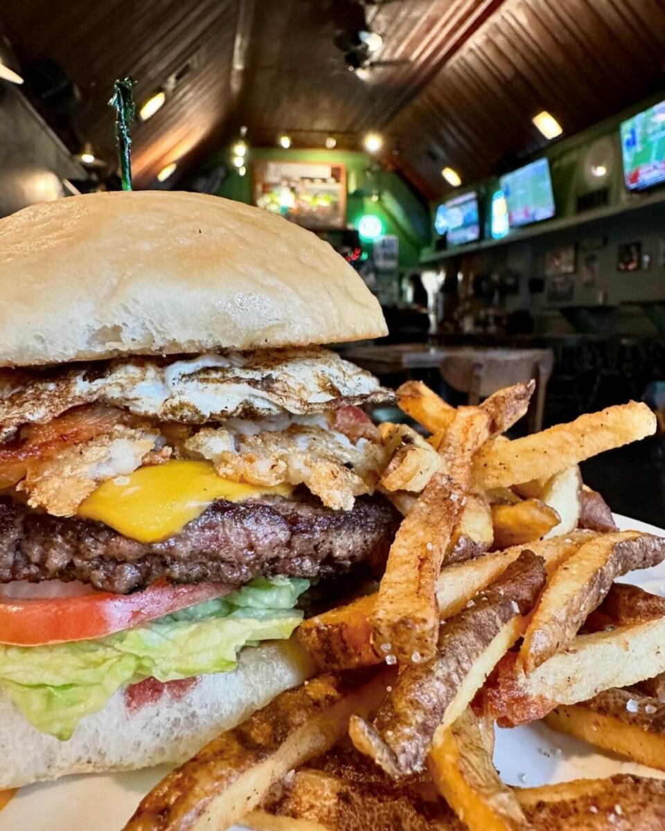 A cheeseburger with lettuce, tomato, bacon, fried egg, and a side of seasoned fries on a plate, inside a restaurant with TVs in the background.