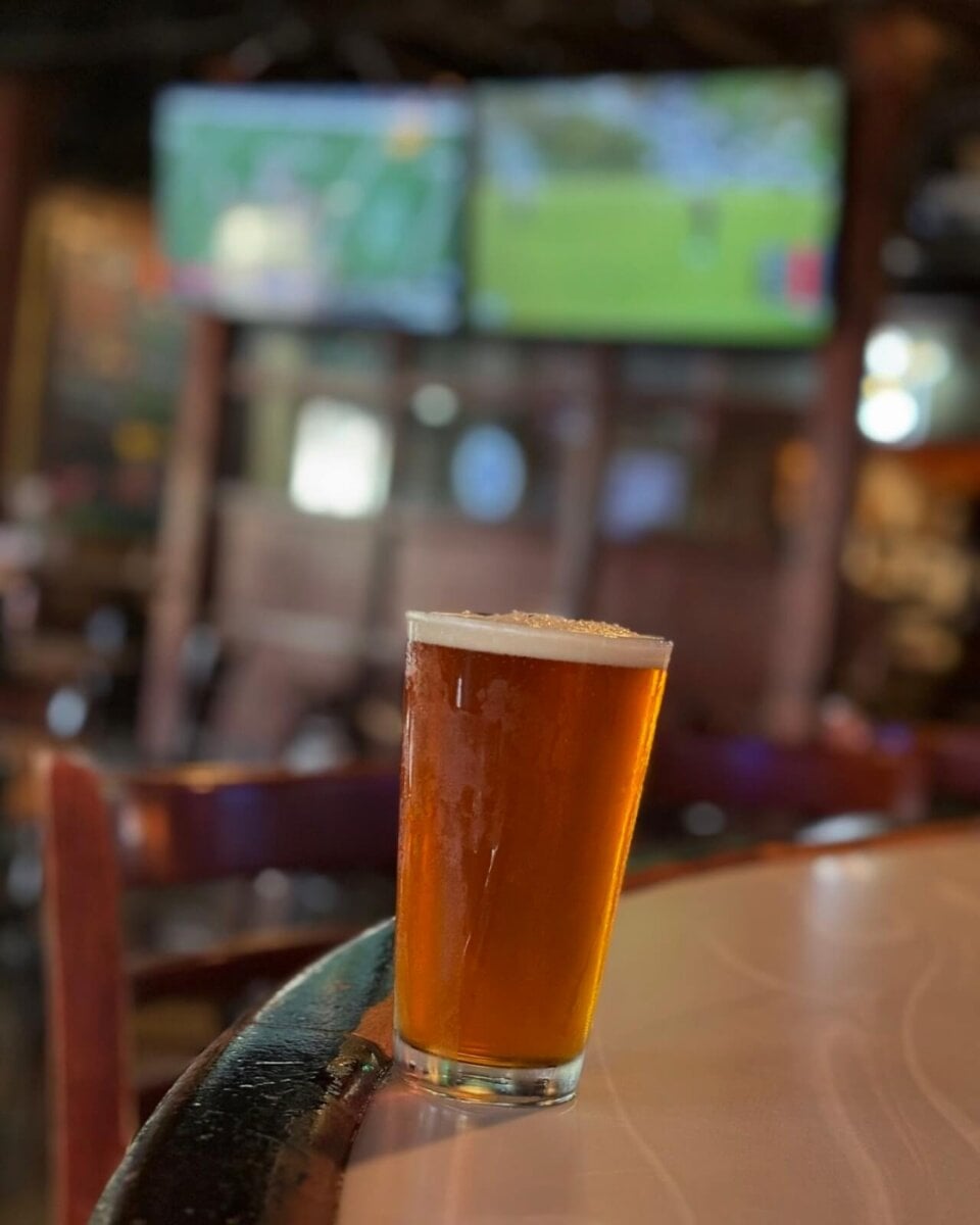 A pint glass of amber beer sits on a bar counter in a dimly lit pub, with two blurred TV screens showing sports in the background.