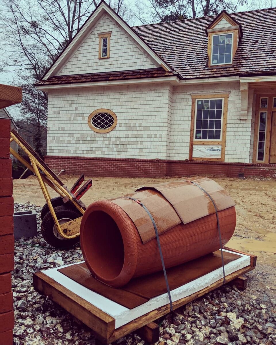 You’re About to Notice Chimney Pots Everywhere - 3 A large terracotta pipe, reminiscent of traditional chimney pots, lies strapped to a pallet in front of a partially constructed house with white siding and a shingled roof.