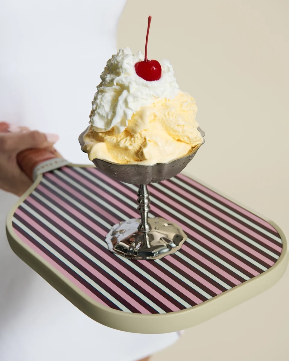 A silver dish of ice cream with whipped cream and a cherry sits on a striped paddle.