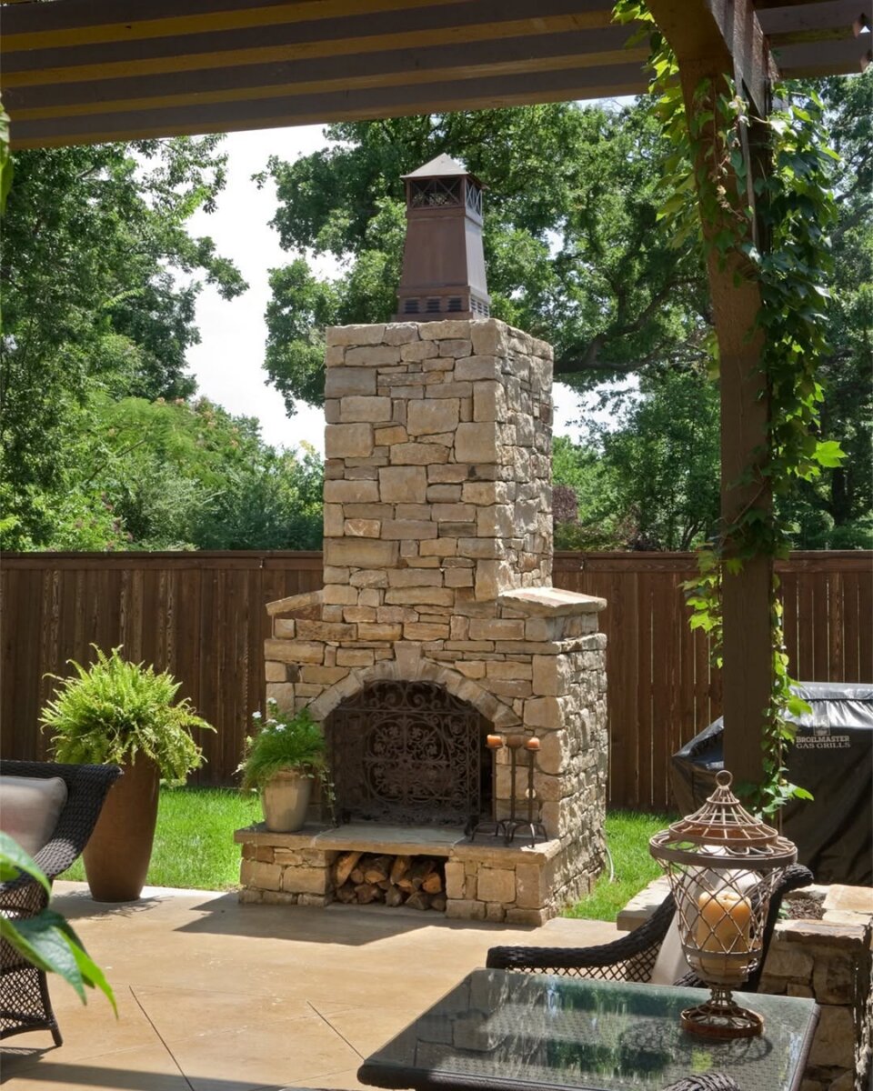 You’re About to Notice Chimney Pots Everywhere - 7 Outdoor stone fireplace with a metal chimney accented by classic chimney pots, surrounded by patio furniture, plants, and a wooden fence, with trees visible in the background.