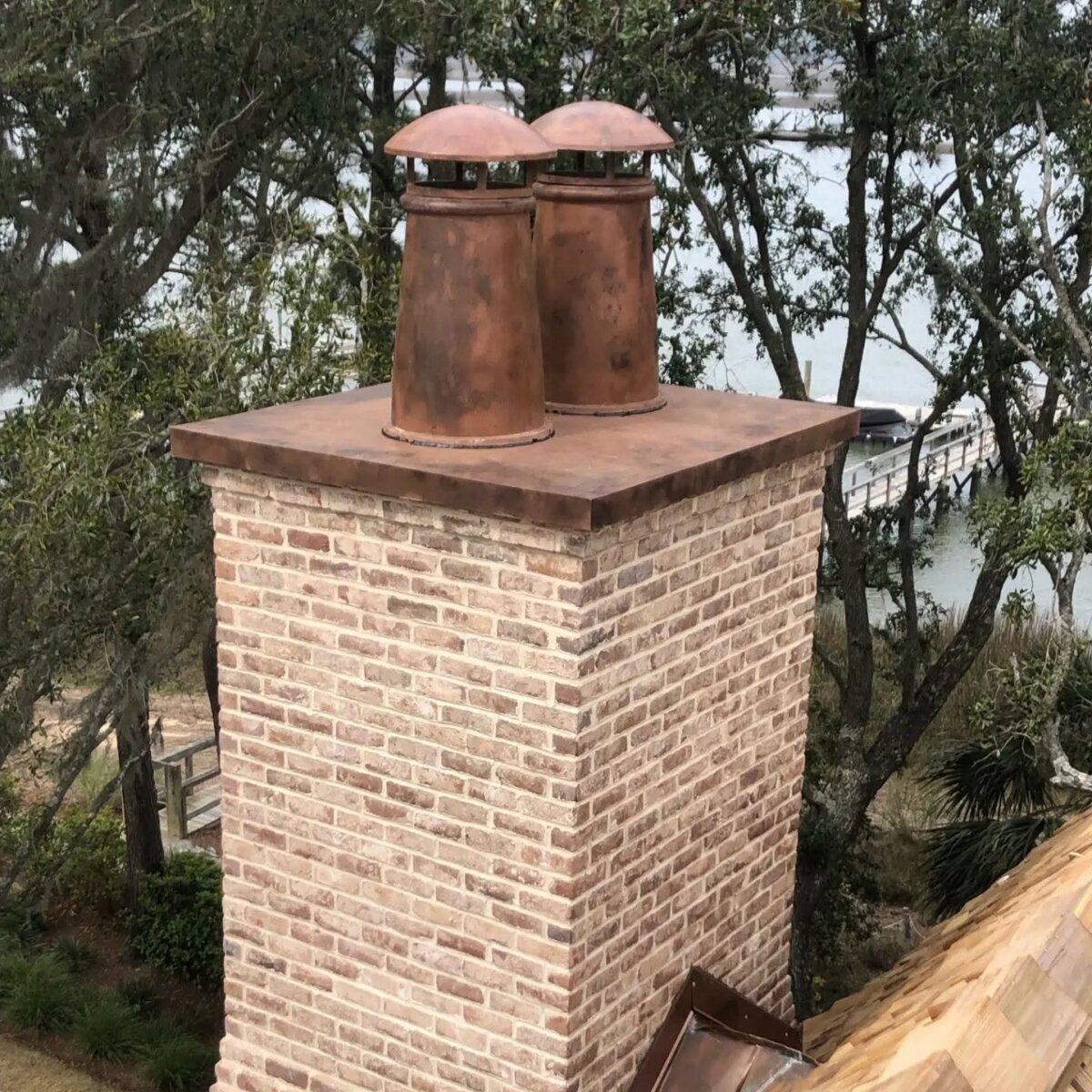 You’re About to Notice Chimney Pots Everywhere - 6 A brick chimney with two rust-colored chimney pots stands on a rooftop, surrounded by trees and overlooking a body of water.