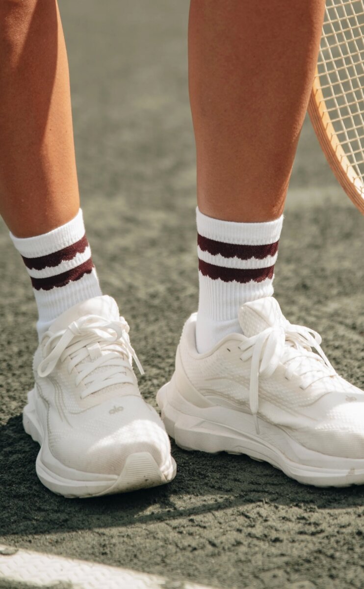 Close-up of a person showcasing effortless court style, wearing white sneakers and white socks with two maroon stripes, standing on a tennis court next to a tennis racket.