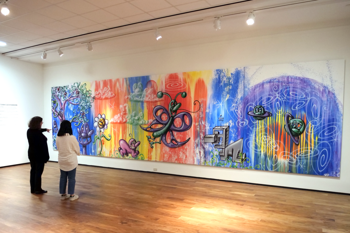 Two people view a large, colorful mural with whimsical animal figures and abstract shapes on display in a mobile art exhibit set within an art gallery with wooden floors.