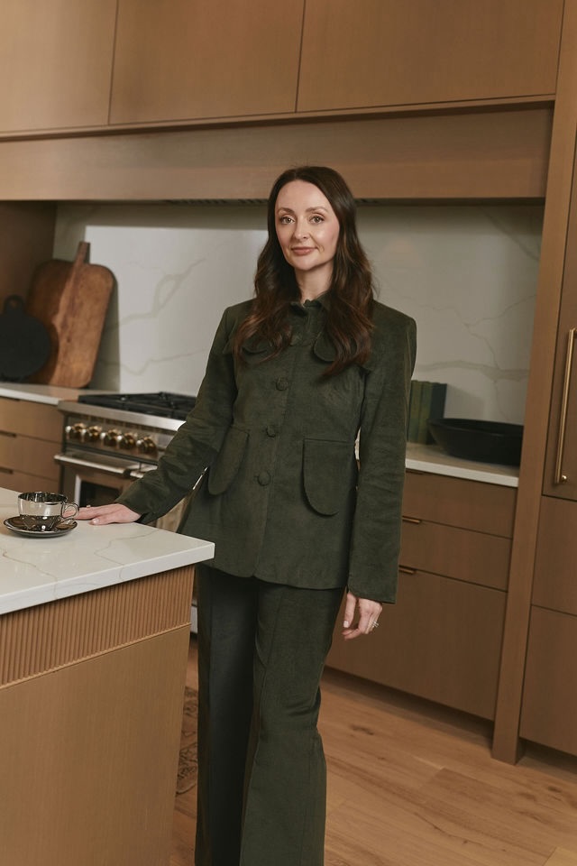 Meet the Sisters Behind Muracha: The South's New Must-Try Matcha - 3 A woman in a dark green suit stands in a modern kitchen with wood cabinets, a marble backsplash, and a stove in the background, thoughtfully preparing a cup of premium matcha.