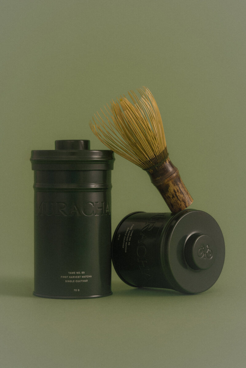 Meet the Sisters Behind Muracha: The South's New Must-Try Matcha - 5 Two matte black tea canisters, one upright and one lying down, with a bamboo matcha whisk resting on top, set against a muted green background.