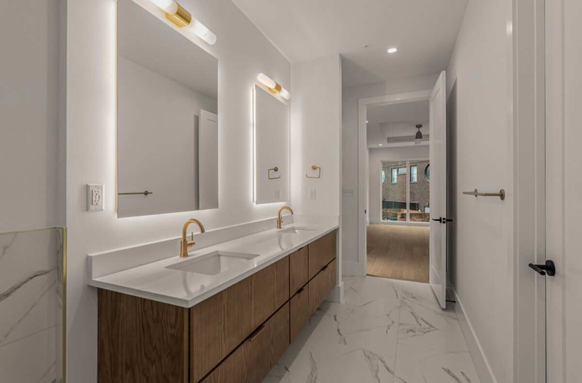 Modern bathroom with double sinks, backlit mirrors, gold fixtures, and a marble floor. A doorway leads to a room with wood flooring and large windows, offering contemporary comfort in the heart of Nashville at 321 31st Ave N.