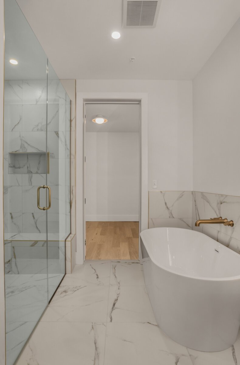 Modern bathroom at 321 31st Ave N, Nashville, featuring a glass-enclosed shower, a white freestanding bathtub, marble-style flooring and walls, and a gold faucet. A doorway leads to a room with wooden flooring in this stunning Nashville TN home.
