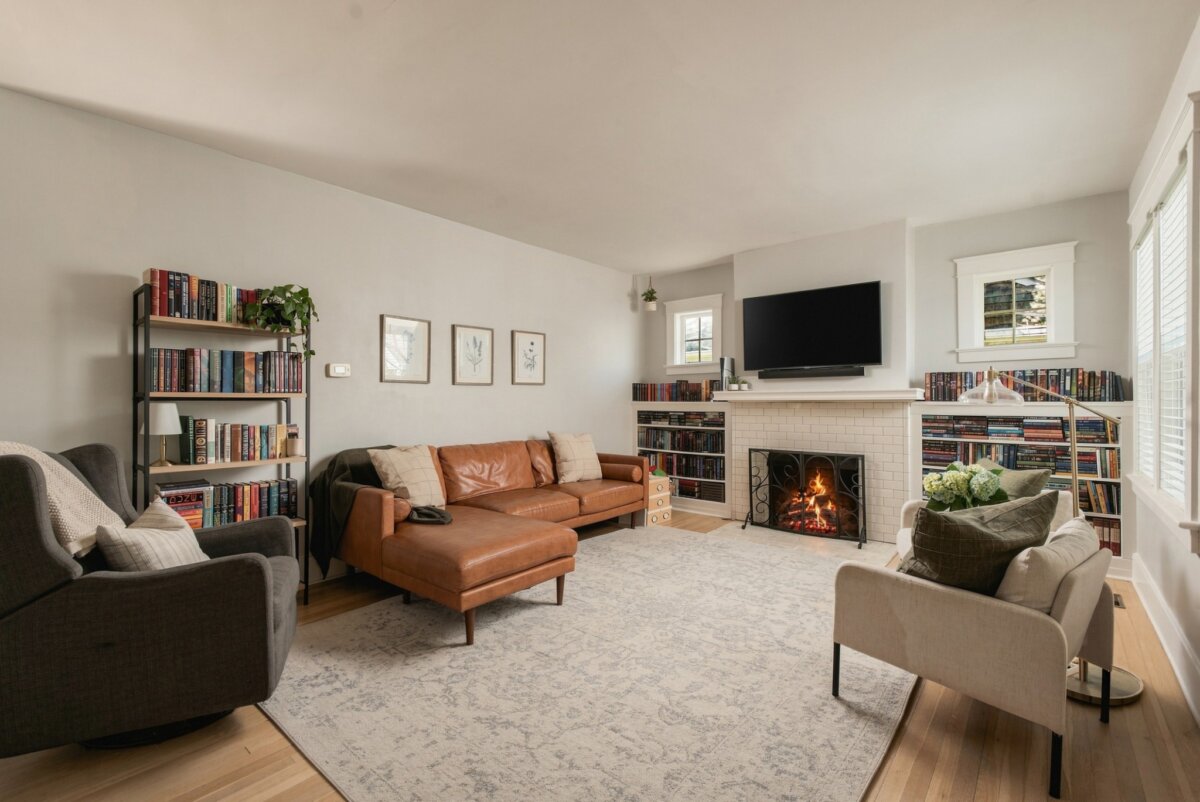 Living room at 2505 W Linden Ave, Nashville, featuring a brown leather sectional, armchair, bookshelves, fireplace with fire, wall-mounted TV, and large area rug. Natural light enters through two windows.