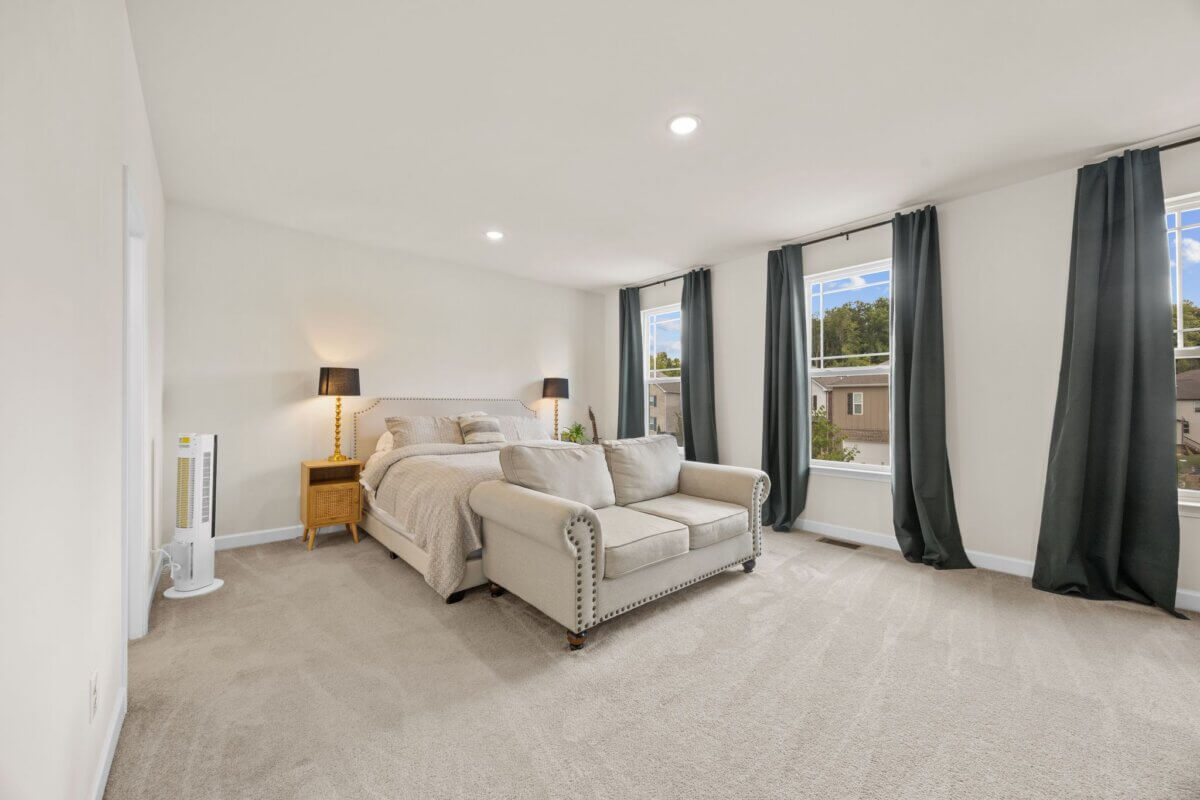 Bright bedroom in Nashville, TN, featuring a comfortable bed with a beige loveseat at the foot, nightstand with lamp, carpeted floor, and three large windows adorned with dark curtains—located at 1121 Parklawn Dr.