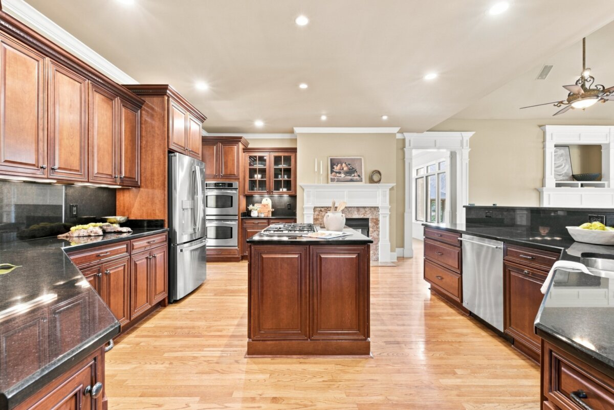Spacious kitchen with wood cabinets, black countertops, stainless steel appliances, center island, hardwood floors, and a fireplace in the background—perfect for entertaining in this beautiful home on Steelson Way in Murfreesboro, TN.