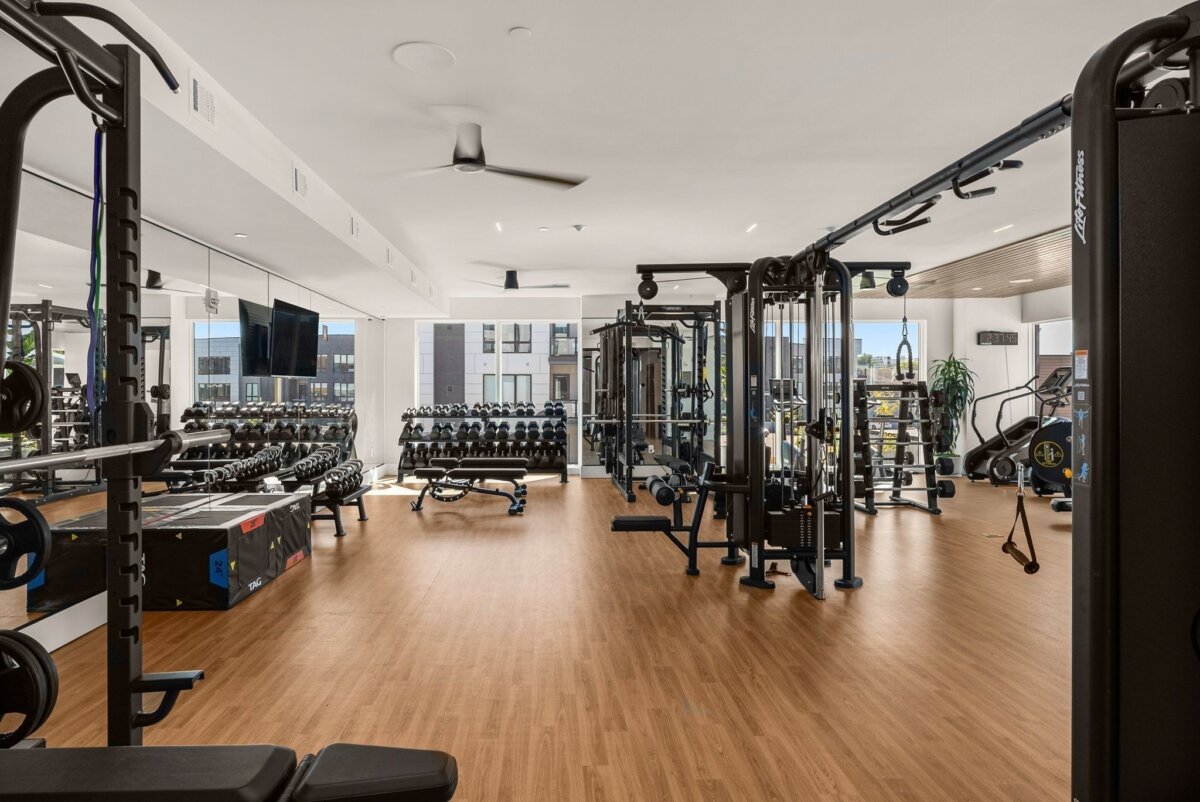 Modern gym with wood flooring featuring free weights, exercise machines, benches, mirrors, and large windows allowing natural light into the space—conveniently located within the apartment community at 321 31st Ave N in Nashville.