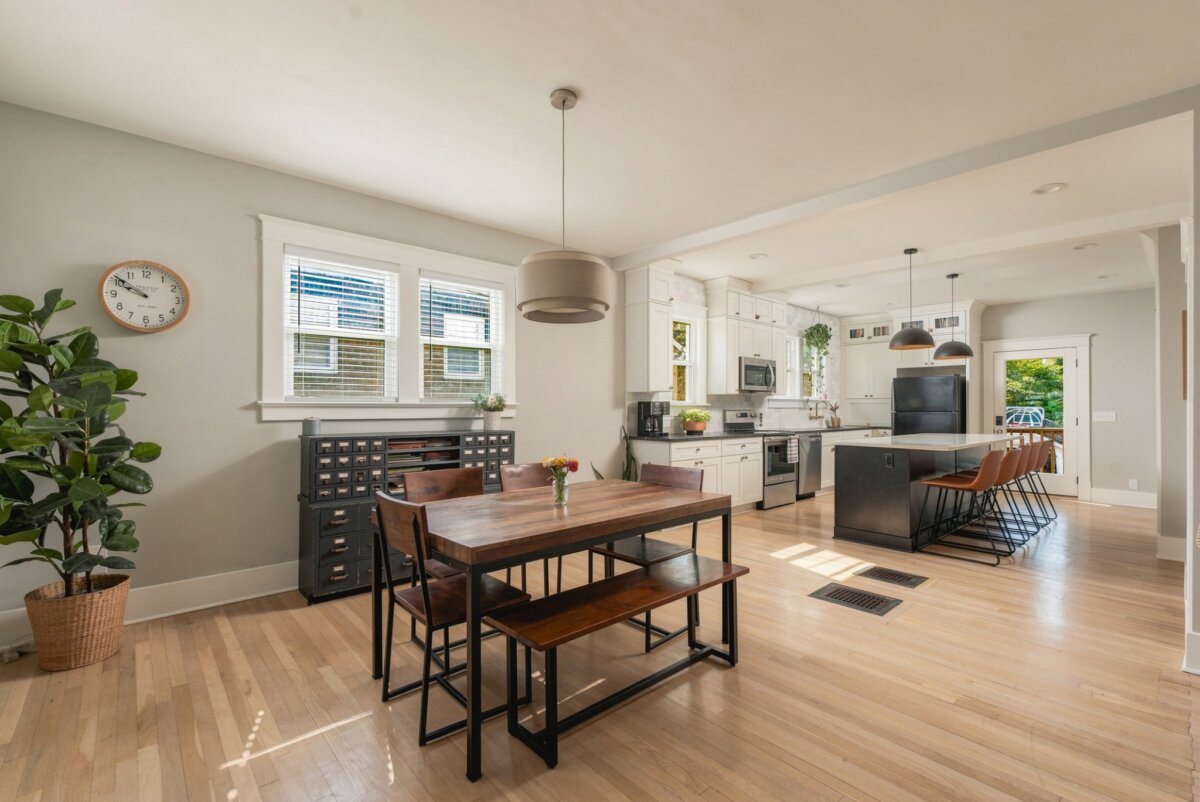 Bright, modern kitchen and dining area at 2505 W Linden Ave, Nashville TN, featuring wood floors, a large table, bar stools at a kitchen island, houseplants, a wall clock, and windows letting in natural light.