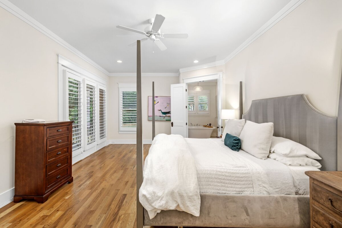 Bright Nashville bedroom with a four-poster bed, hardwood floors, a wooden dresser, large windows with shutters, and an open door leading to a bathroom at 4000 Wallace Lane.