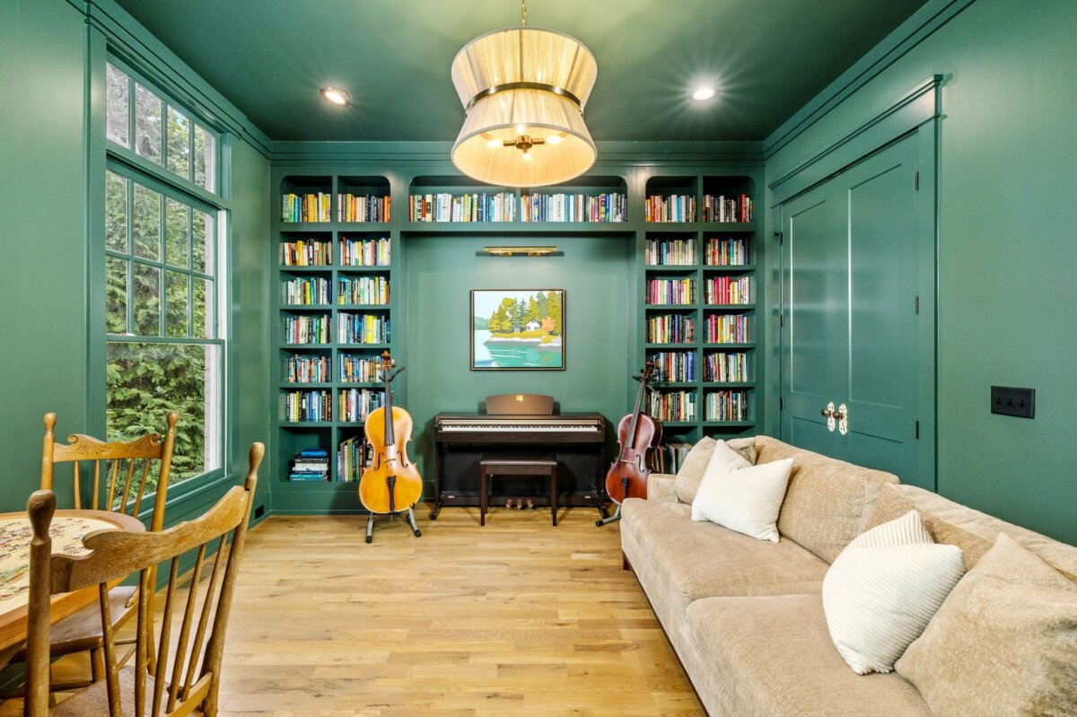 A cozy green-walled room at 4000 Wallace Lane in Nashville, featuring bookshelves, a piano, two string instruments, a sofa, a wooden dining table, large windows, and a modern chandelier.