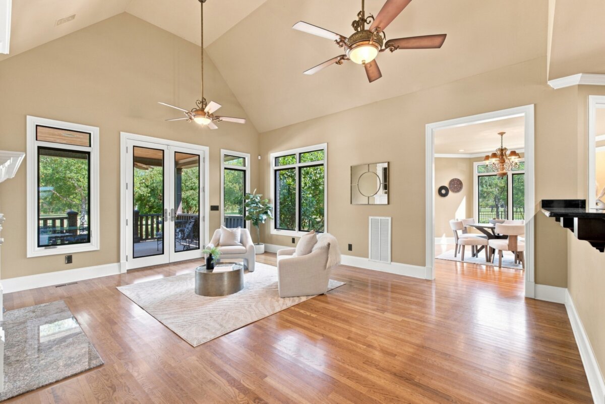 Spacious living room with hardwood floors, beige walls, two armchairs on a rug, ceiling fans, and large windows leading to a covered patio at 218 Steelson Way. Dining area visible through an open doorway in this inviting Murfreesboro, TN home.