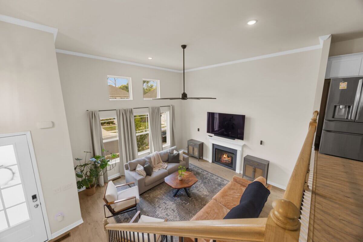 Modern living room with large windows, neutral walls, a ceiling fan, a TV above a fireplace, couches, a round coffee table, and a potted plant near the window—all beautifully arranged in this stunning home at 1121 Parklawn Dr in Nashville TN—viewed from the staircase.