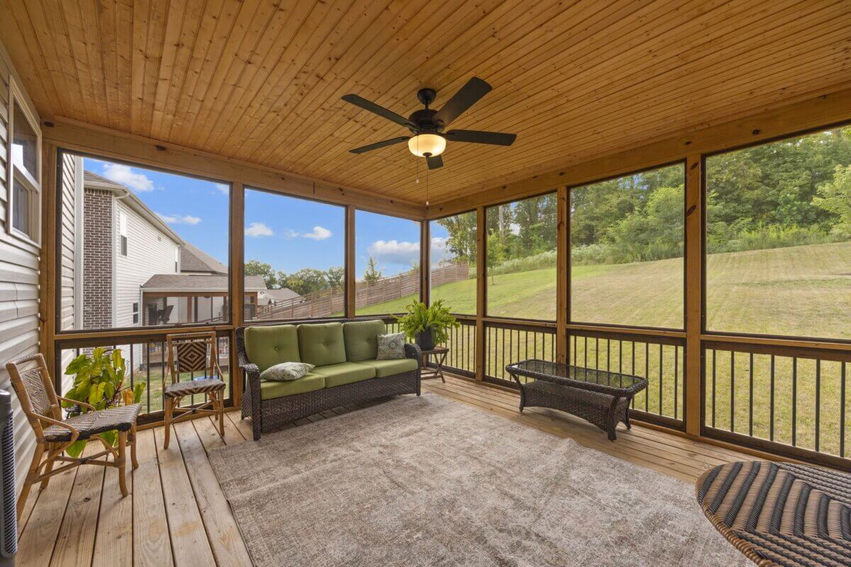 Screened porch with wooden floor and ceiling, ceiling fan, wicker furniture, rug, potted plants, and large windows overlooking a grassy backyard and neighboring houses in Nashville TN. This inviting outdoor space at 1121 Parklawn Dr offers comfort and relaxation in a charming setting.