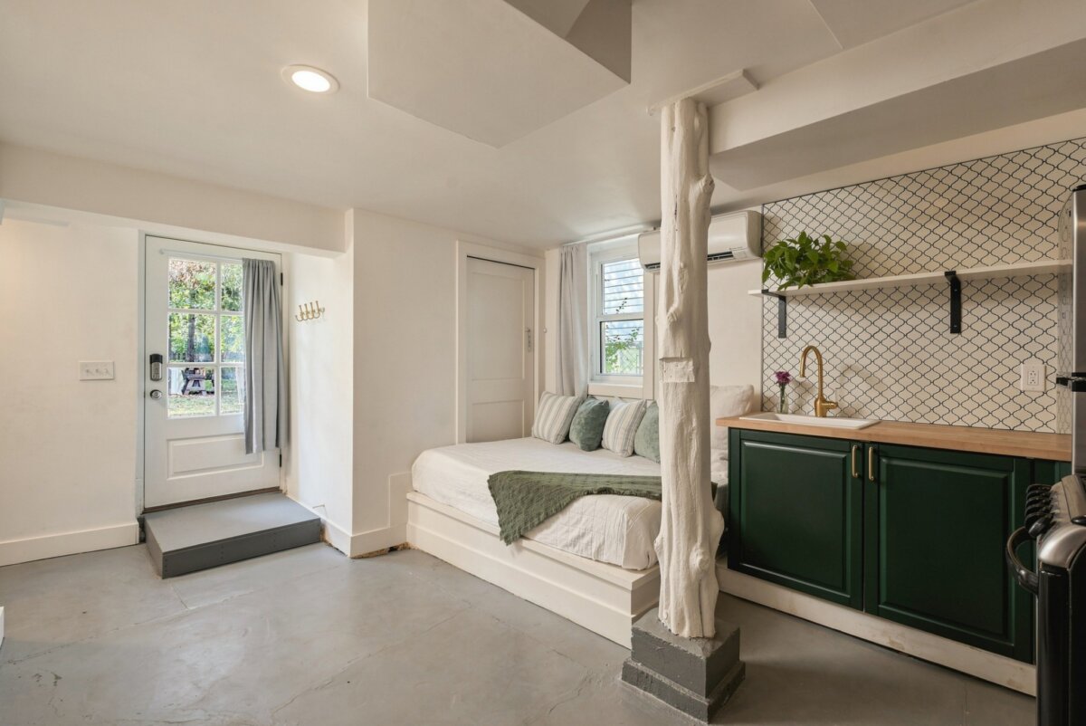 A small, modern studio located at 2505 W Linden Ave in Nashville, TN, featuring a bed, green cabinets, a kitchenette, white walls, and a door leading outside.