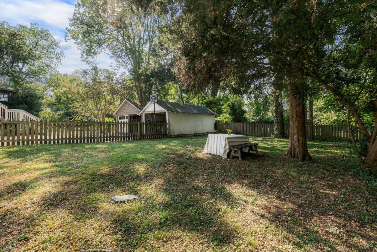 A backyard with scattered shade, a wooden fence, a shed, trees, and a picnic table covered with a striped cloth creates a charming retreat at 2505 W Linden Ave in Nashville, TN.
