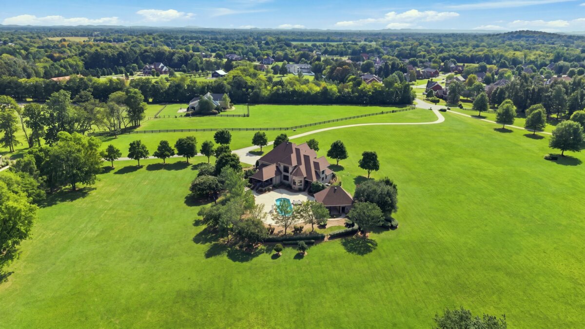 Aerial view of a large house with a pool on 218 Steelson Way, surrounded by trees and open green fields, with neighboring homes in the distance in Murfreesboro, TN.