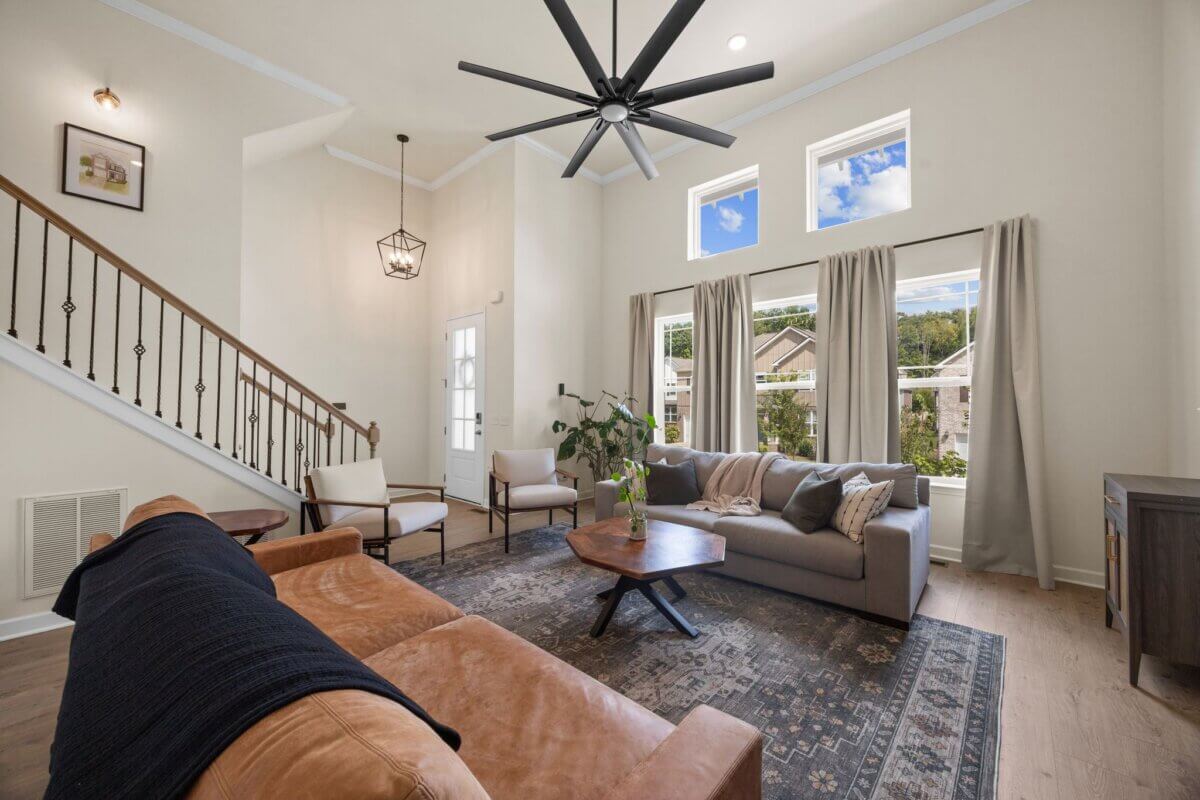 Modern living room at 1121 Parklawn Dr, Nashville TN, featuring a brown leather sofa, gray couch, two chairs, large ceiling fan, three windows with curtains, and a staircase with a wooden railing.