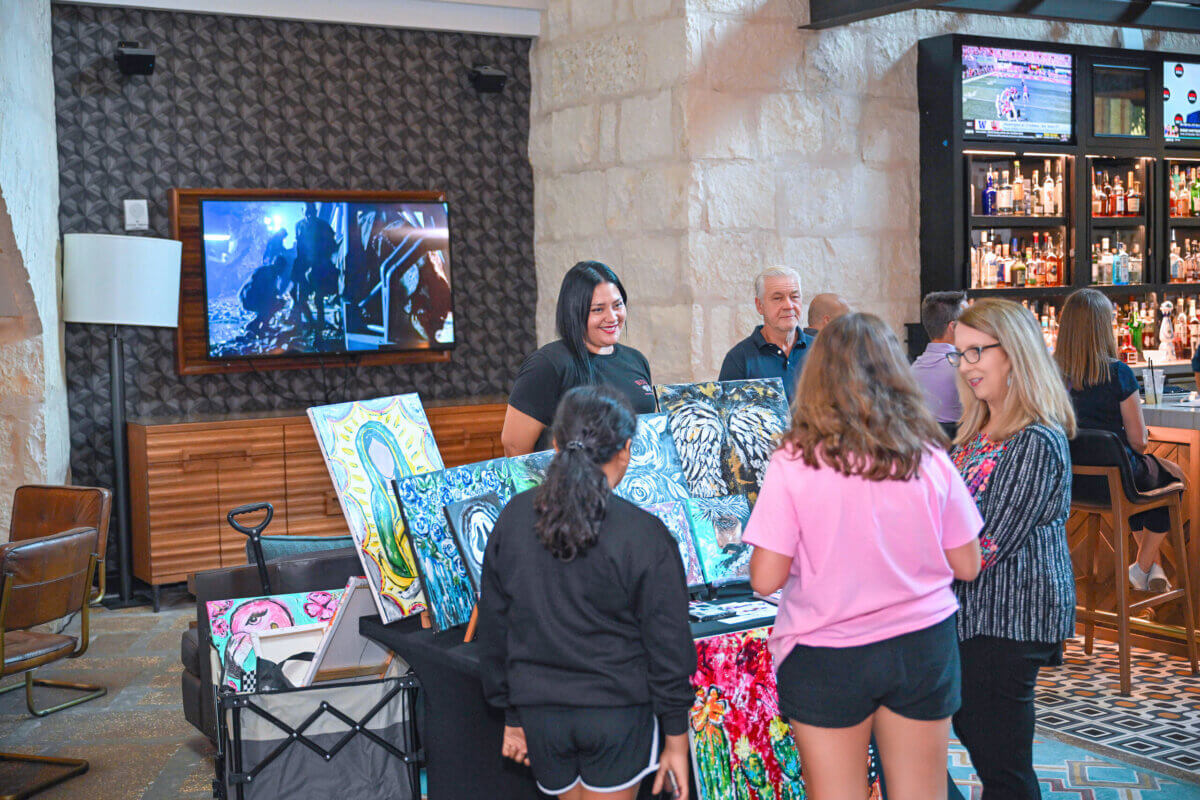 A group of people views colorful paintings displayed on a table inside a bar or lounge area, with TVs and a bar visible in the background—a unique blend of art and travel that highlights clever hotel amenities designed to enrich every guest’s stay.