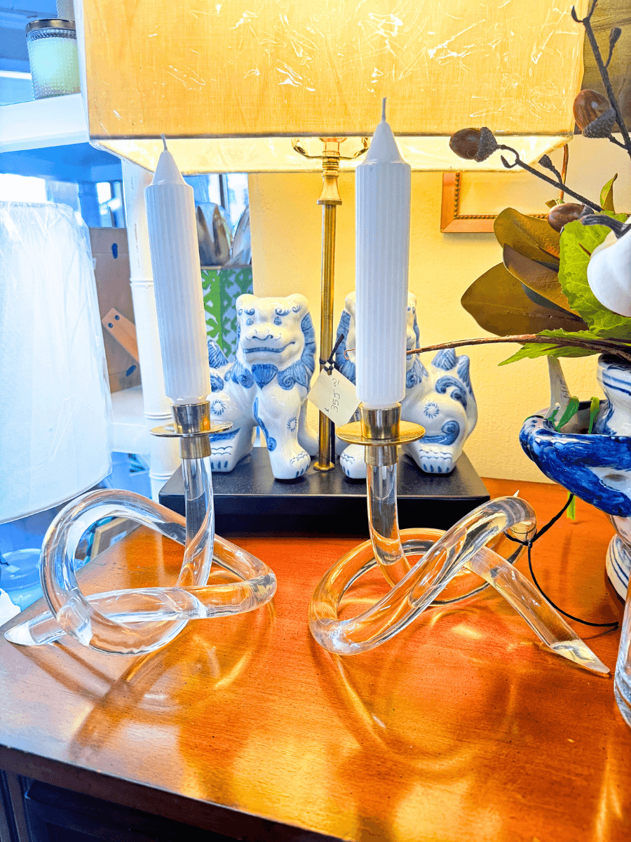 17 Louisville FINDS for October: Fall Fashion, Festive Decor & Game-Day Essentials - 13 Two clear glass candlesticks with white candles add to the cozy finds on a wooden table in front of a lamp and blue-and-white ceramic lion figurines.