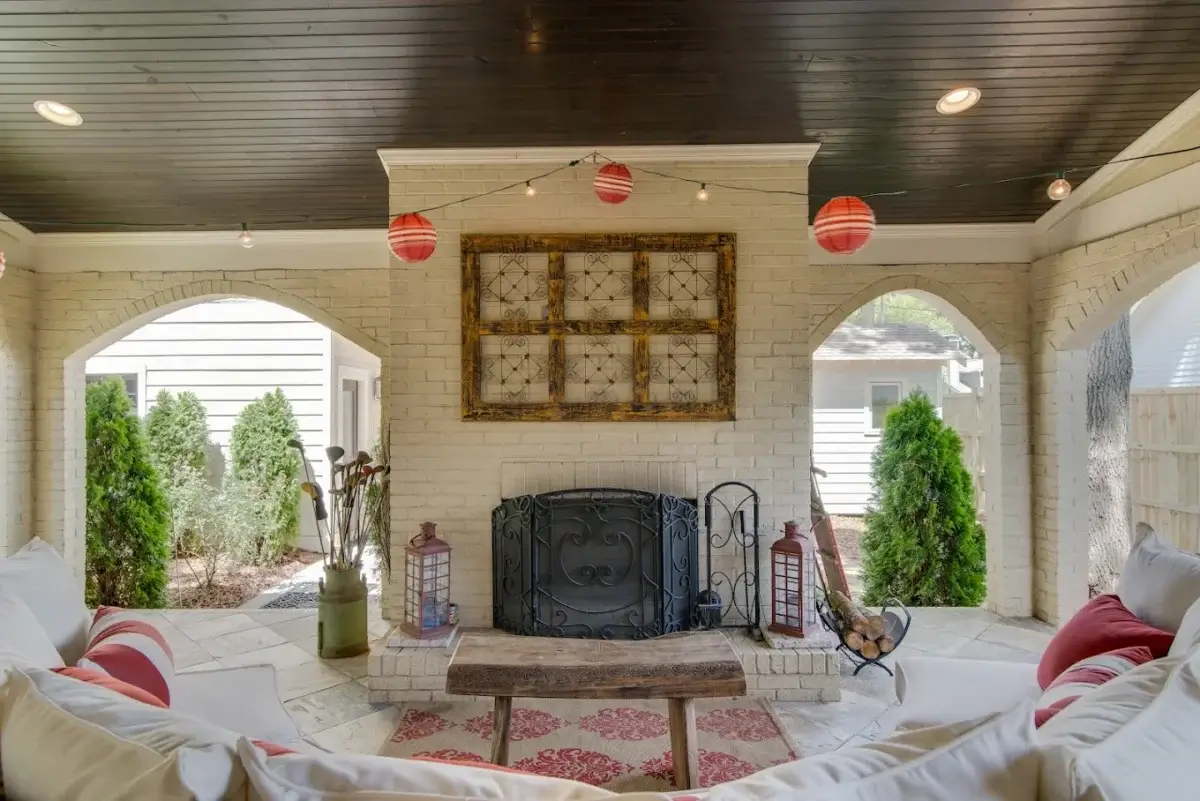 Covered patio with white brick walls, a fireplace, a wooden bench, string lights, red paper lanterns, and cushioned seating with red accents. Shrubs and lanterns decorate the space, creating an inviting atmosphere reminiscent of the stylish gathering spaces found in Nashville boutique hotels.