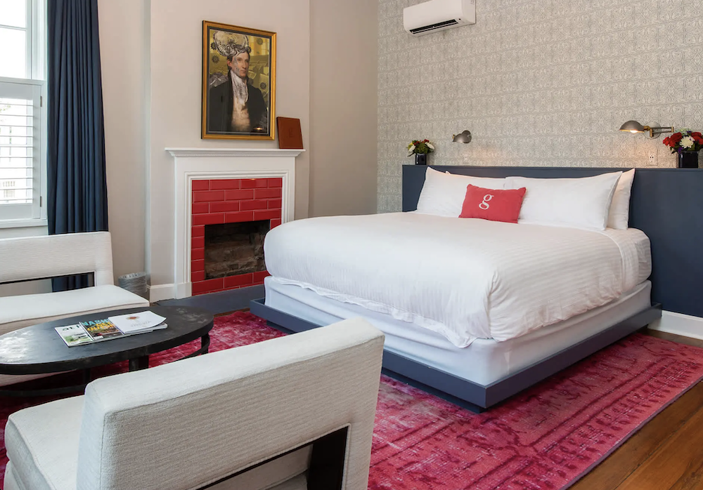 5 Nashville Boutique Hotels to Buy Out for Your Wedding Weekend - 3 Modern boutique hotel room in Nashville featuring a white bed with a red accent pillow, fireplace with red tiles, art above the mantel, two white chairs, round table, and a bright pink rug—perfect for a memorable wedding weekend.
