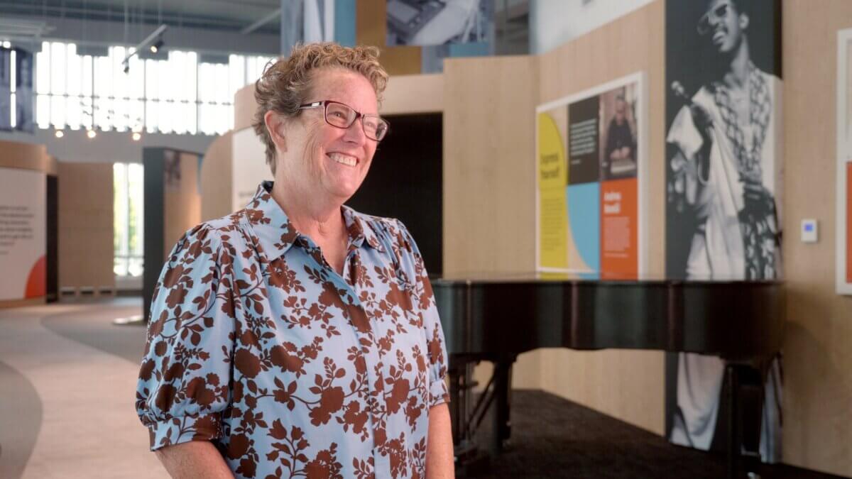 She's Making the Blind Feel Seen Through Louisville’s Newest Museum Experience - 2 A person wearing glasses and a floral shirt stands smiling in a modern Louisville museum exhibition space, with display panels and a piano in the background, capturing the essence of an engaging museum experience.