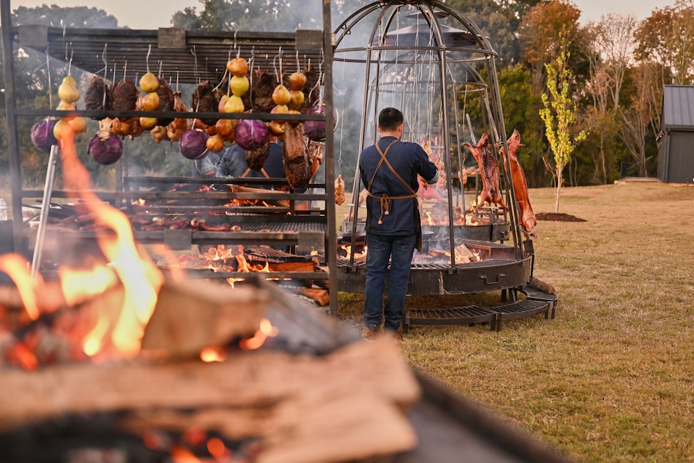 A person practices live-fire cooking, preparing meat and vegetables over an open fire at an outdoor barbecue setup, with food hanging and roasting on metal frames.