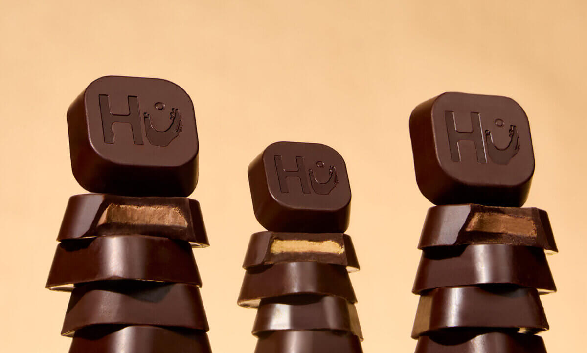 10 Grown-Up Halloween Candies We’re Keeping for Ourselves - 3 Three stacks of dark chocolate pieces, each topped with a piece engraved with the “Hu” logo, are arranged against a beige background—offering a grown-up guide to gourmet Halloween candy for those seeking an elevated treat this season.