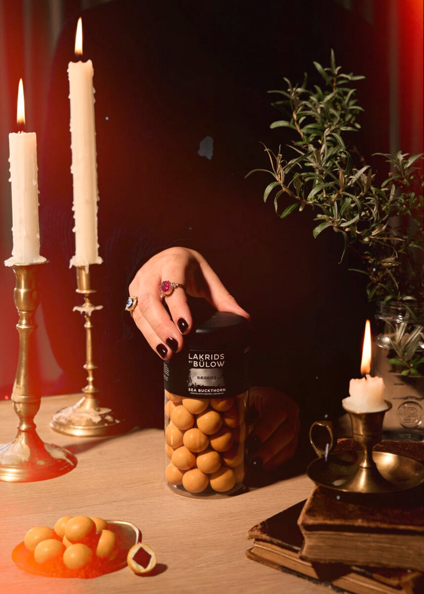 10 Grown-Up Halloween Candies We’re Keeping for Ourselves - 2 A hand with dark nail polish holds a jar of Lakrids by Bülow—a true grown-up Halloween candy—on a table adorned with candles, greenery, books, and some round gourmet candies.