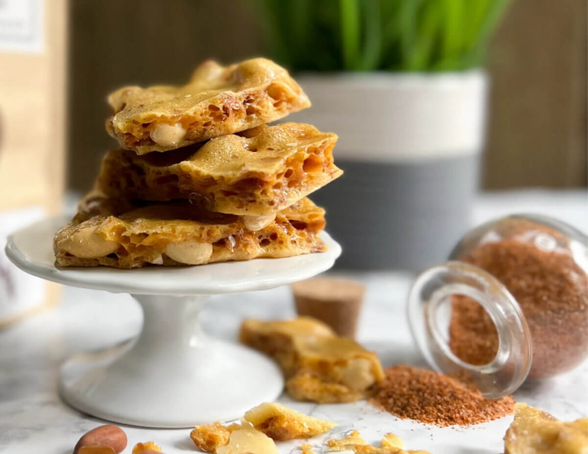 10 Grown-Up Halloween Candies We’re Keeping for Ourselves - 11 Pieces of peanut brittle stacked on a small white cake stand, with spilled sugar and nuts nearby, and a plant in the background—an elegant display perfect for any grown-up guide to gourmet Halloween candy.