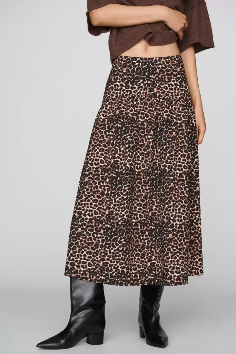 Fall Is Getting Wild: Animal Prints Are Back! - 12 A person wearing a brown top, a leopard print tiered skirt, and black knee-high boots stands against a plain background, showcasing one of this season’s standout autumn trends: animal prints in fall fashion.