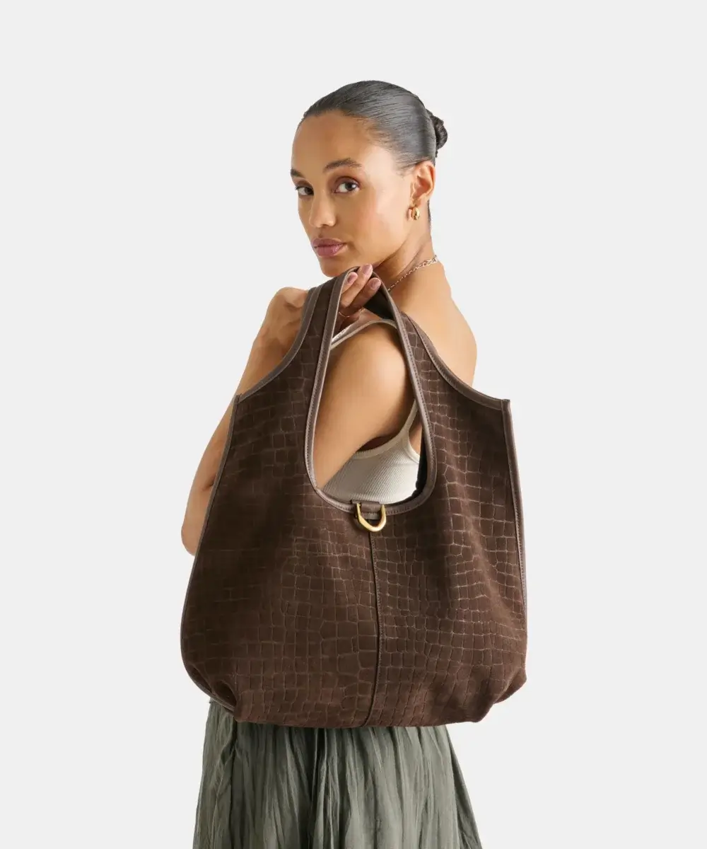 Fall Is Getting Wild: Animal Prints Are Back! - 23 Woman facing sideways with a brown textured shoulder bag, wearing a sleeveless top and pleated skirt featuring subtle animal print trends, against a plain light background.