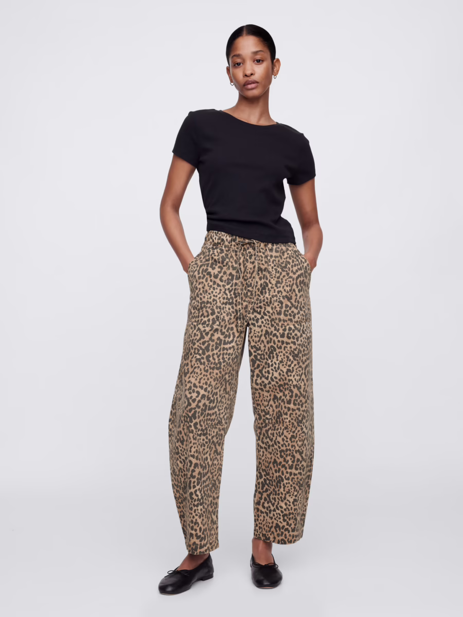 Fall Is Getting Wild: Animal Prints Are Back! - 14 Person standing against a plain background, wearing a black short-sleeve top, loose-fitting leopard-print pants that showcase the latest in Animal Print Trends, and black shoes, with hands in pockets.