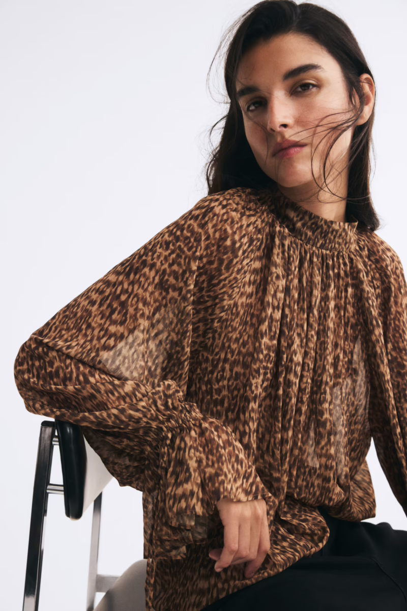 Fall Is Getting Wild: Animal Prints Are Back! - 3 Person sitting on a chair wearing a sheer, long-sleeve, leopard print blouse with pleated details—a chic nod to animal print trends perfect for elevating your fall fashion wardrobe.