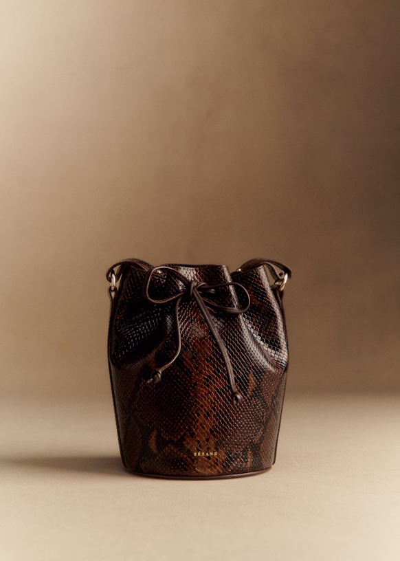 Fall Is Getting Wild: Animal Prints Are Back! - 21 A brown, snakeskin-textured bucket bag with a drawstring closure—perfect for fall fashion—sits upright against a neutral beige background, adding a touch of bold animal prints to your seasonal wardrobe.