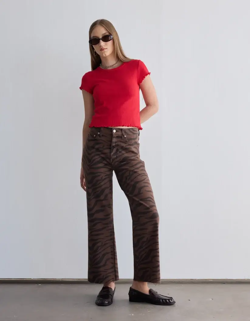 Fall Is Getting Wild: Animal Prints Are Back! - 13 Person wearing a red short-sleeve top, brown tiger-striped wide-leg pants inspired by Animal Print Trends, black loafers, and sunglasses stands against a plain white wall.