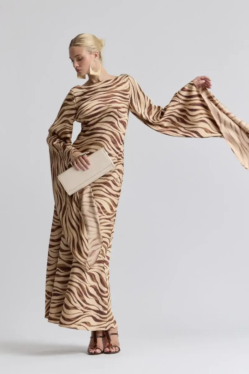 Fall Is Getting Wild: Animal Prints Are Back! - 5 Person wearing a long, flowing, zebra-print dress with flared sleeves—a perfect nod to Animal Prints—holding a beige clutch bag and wearing strappy sandals, stands against a plain light background.