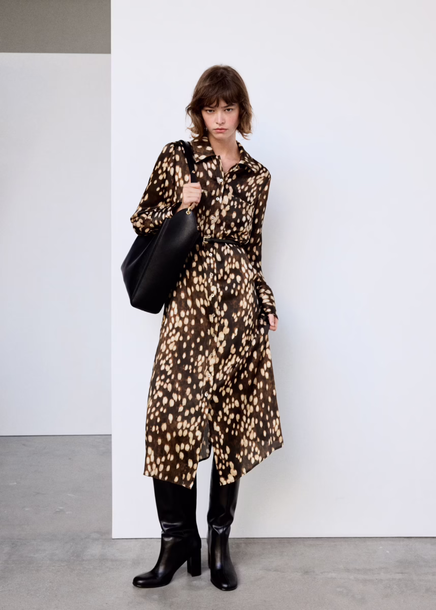 Fall Is Getting Wild: Animal Prints Are Back! - 7 Person standing indoors wearing a brown animal print dress, black boots, and holding a large black bag, with a neutral white and gray background—an effortlessly chic look that captures the essence of fall fashion.