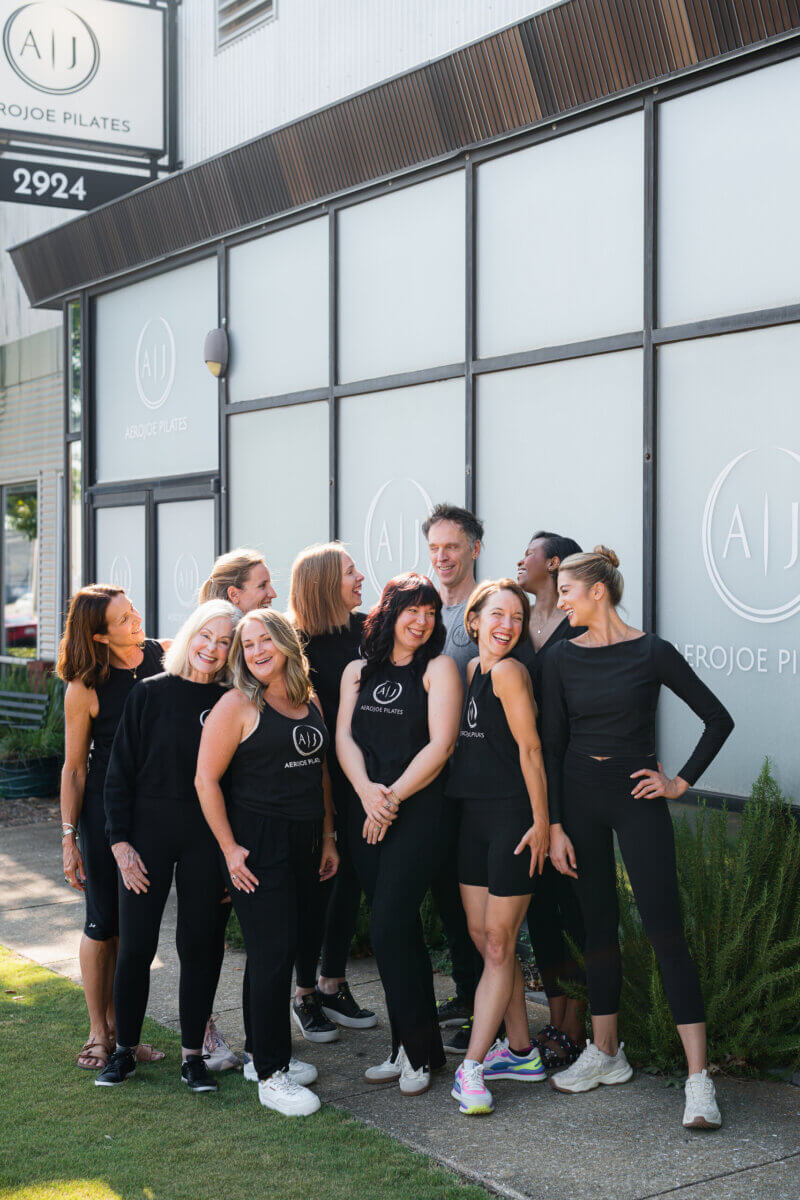 Pilates: Is It Stretching, Strength Training, or Both? - 7 A group of people in black workout clothes stand and smile together outside a building with “Aerojoe Pilates” signage, radiating strength and camaraderie as they embrace the benefits of Pilates practice.