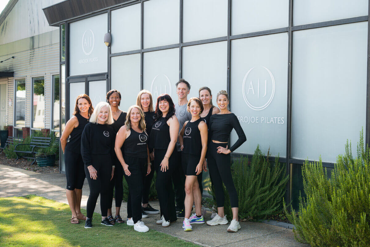 Pilates: Is It Stretching, Strength Training, or Both? - 6 A group of ten adults, dressed in black athletic wear, pose and smile in front of a building labeled “Aero Joe Pilates,” ready to stretch and show off their Pilates strength.