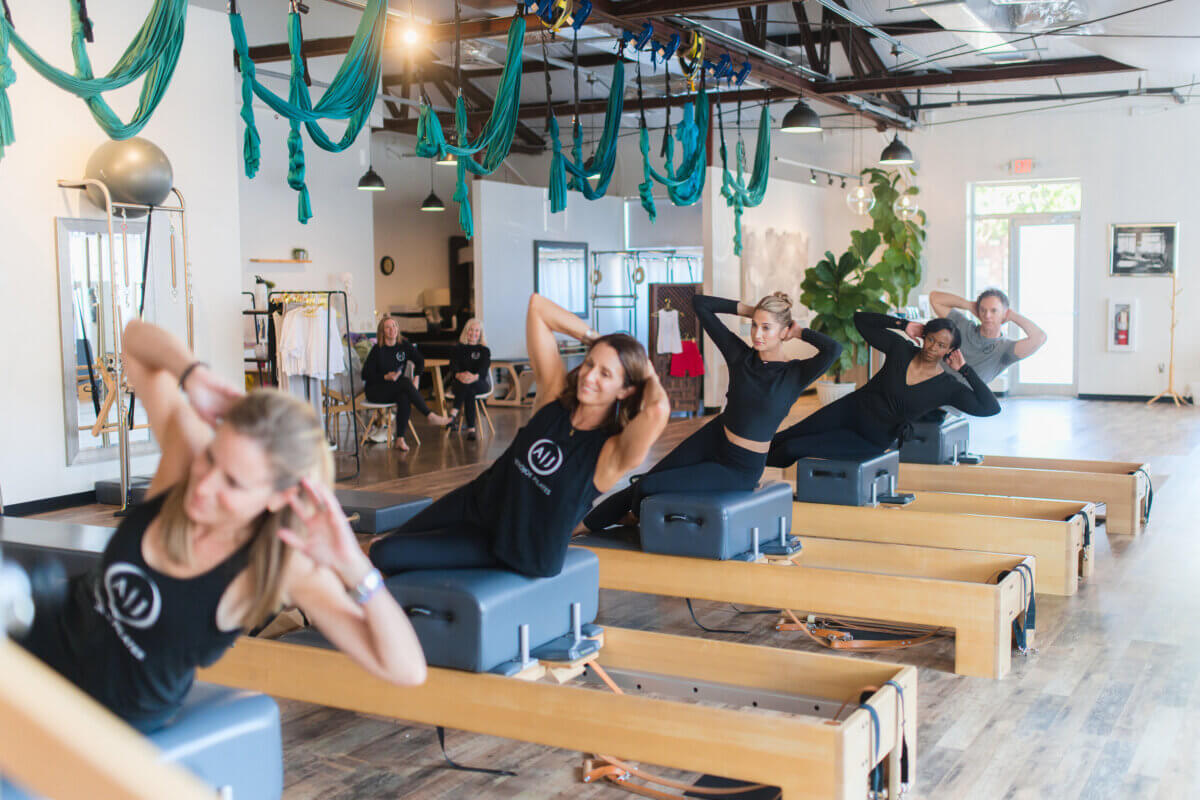 Pilates: Is It Stretching, Strength Training, or Both? - 4 Five women exercise on Pilates reformer machines in a bright studio, performing strength-building abdominal workouts and stretches while an instructor observes in the background.