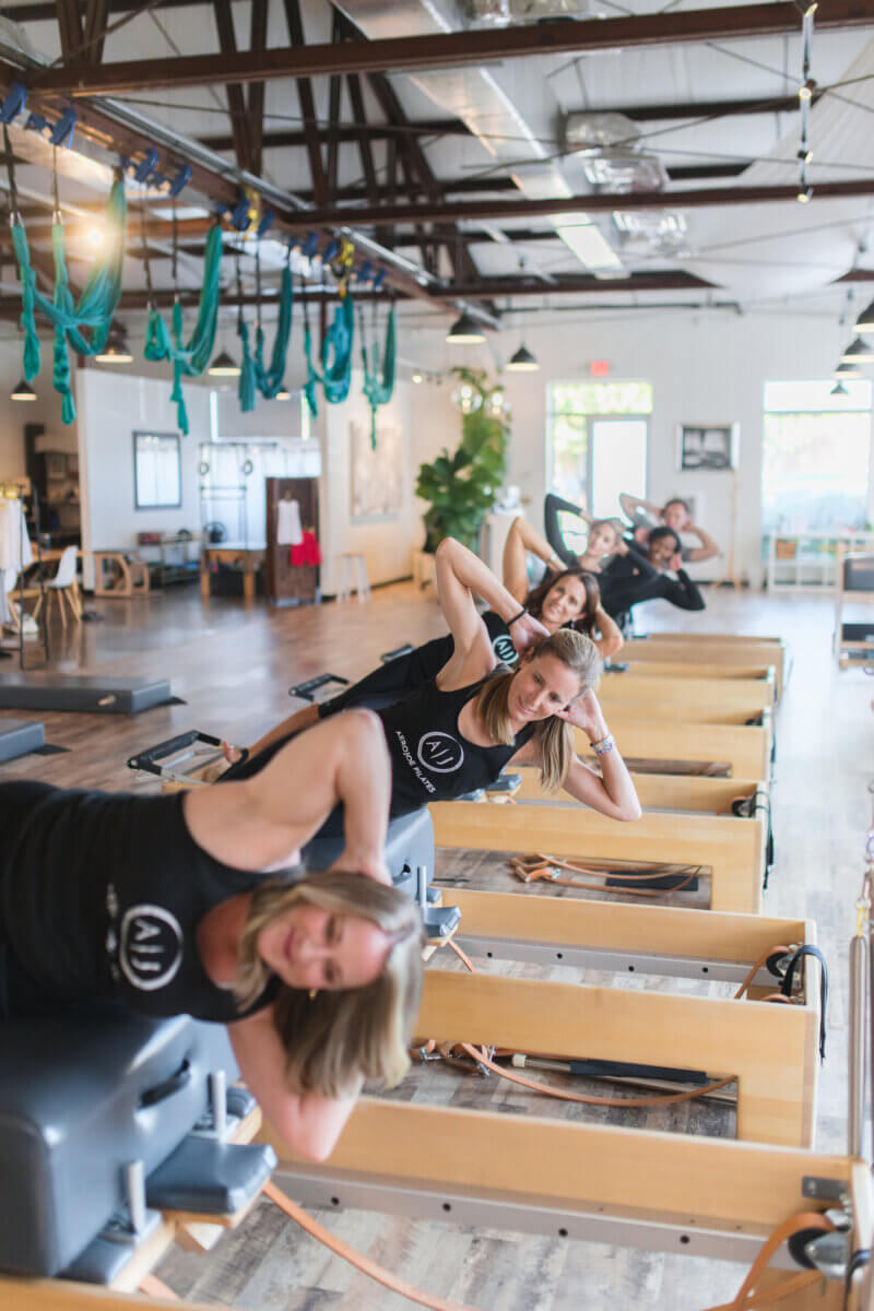 Pilates: Is It Stretching, Strength Training, or Both? - 3 A group of women exercise on Pilates reformer machines in a brightly lit studio with wooden floors and exposed beams, focusing on strength and stretch through their Pilates routines.