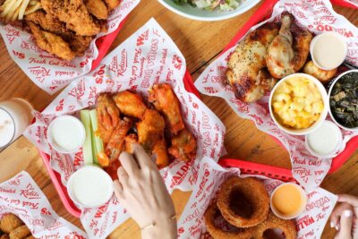 A wooden table showcasing food from new restaurants in Birmingham, featuring red baskets of fried chicken wings, roasted chicken, mac and cheese, onion rings, dipping sauce, and celery sticks.